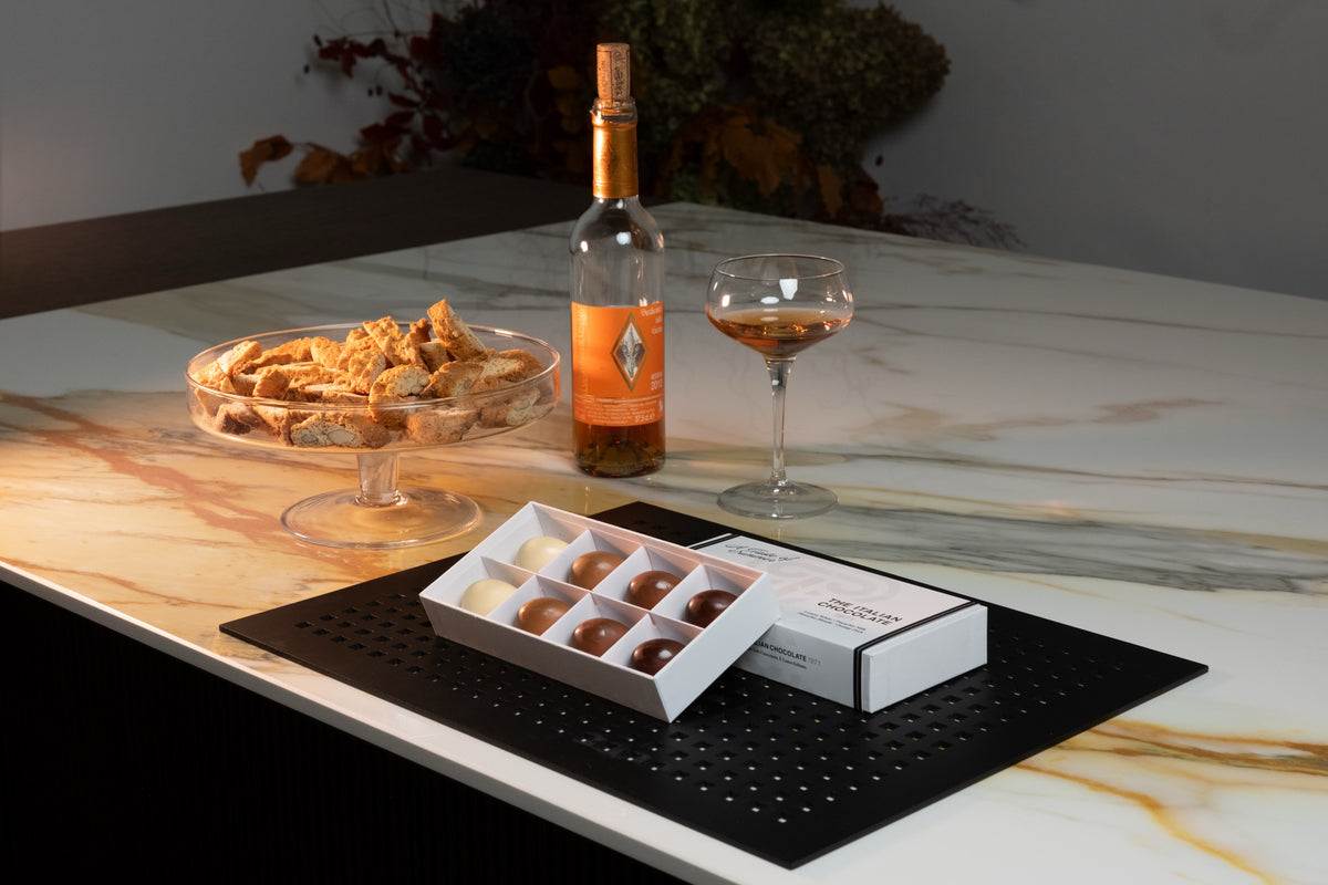 A Taste of Summer box of 8 pralines on a marble table with cantucci and a glass of Vin Santo