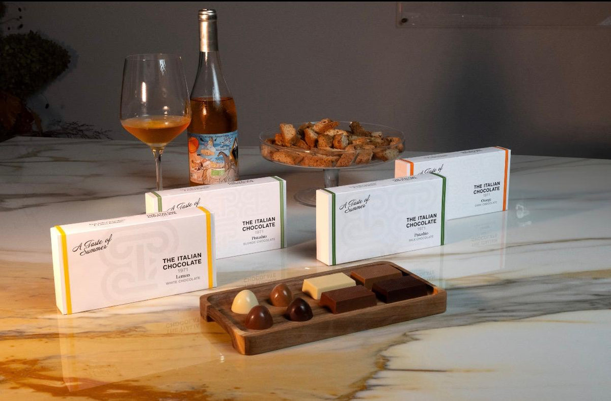 A Taste of Summer chocolate boxes, Lemon, Pistachio and Orange, with pralines and bars on a walnut tray, cantucci and a glass of wine on a marble table